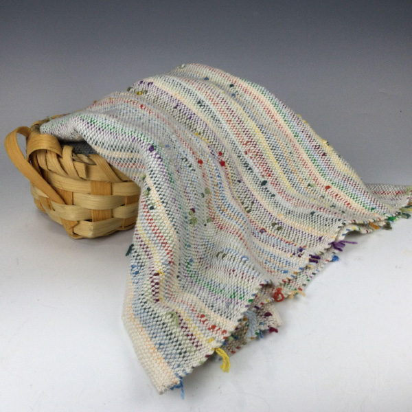 Image of a handcrafted kitchen towel that was woven on a loom as part of the artist's version of Zanshi weaving. The towel is draped inside and over the side of a smaller woven basket as is a mix of white, yellow, blue, red, green, purple, and orange thread. The pattern is woven and textured.