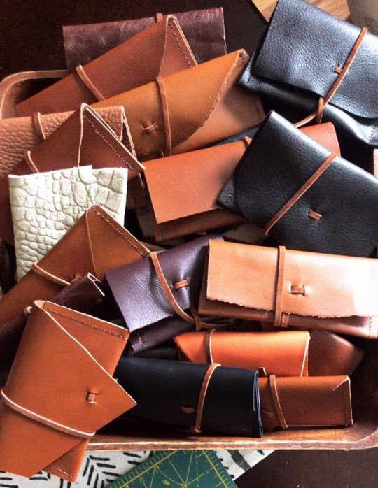 Close up image of seventeen rectangular leather wallets inside of a wooden basket. The colors include light brown, dark brown, white, black, and dark purple. Each wallet has a thin strap wrapped around it.
